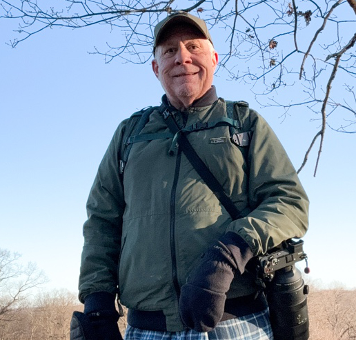 Person outdoors in a jacket and cap, carrying a camera with a long lens. Leafless branches and a clear sky are in the background.